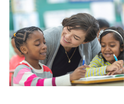 A teacher smiling at two elementary school students working at their desks.