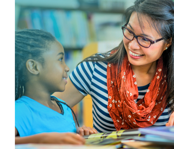 An elementary school student and their teacher sitting at a table next to each other in the library smiling