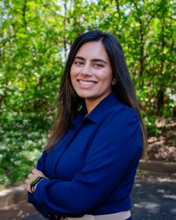 Raheela Charania stands smiling with her arms crosses in front of some green trees. 