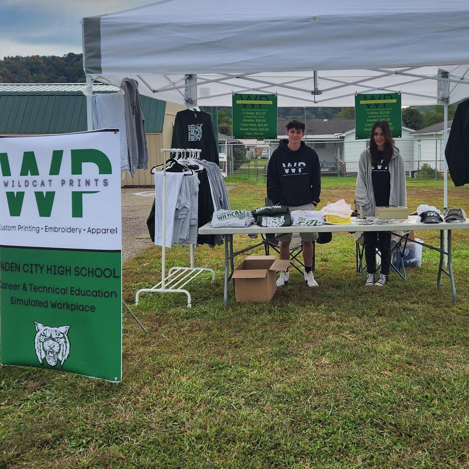 A group of students stand at an outdoor booth, selling T-shirts in front of a Wildcat Prints sign.