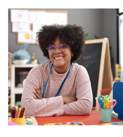 A young teacher sitting at her desk in her classroom. She is wearing a pink sweater and glasses. On top of her desk are cups of colorful school supplies, such as scissors and markers.