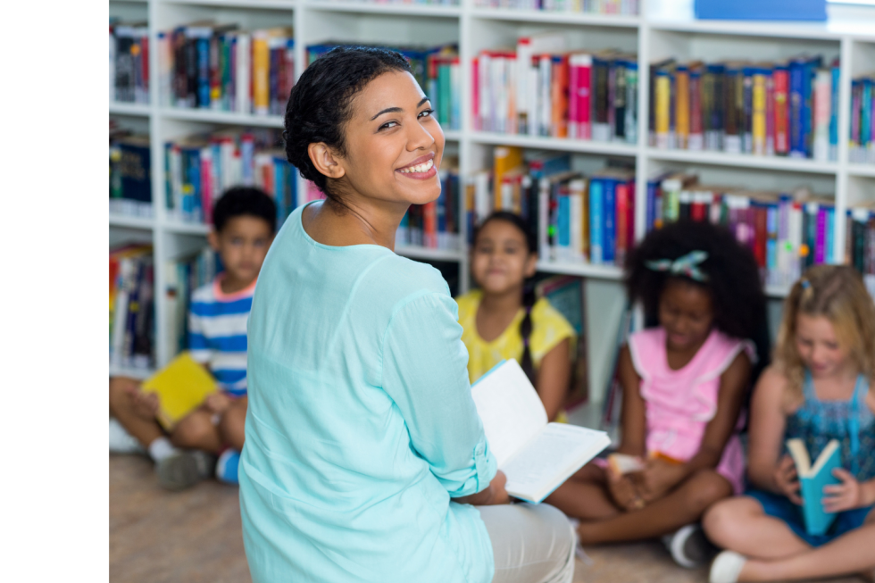 A teacher reading with a group of students in front of some bookshelves.