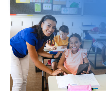 A teacher in a blue shirt supports a smiling student who is using a tablet and has a workbook open on their desk.  Two more students are in the background, focused on their papers in a bright, colorful classroom.