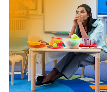 A teacher sitting at a child-size table. Since the pandemic, vacancies are up and retention and applicants are down.
