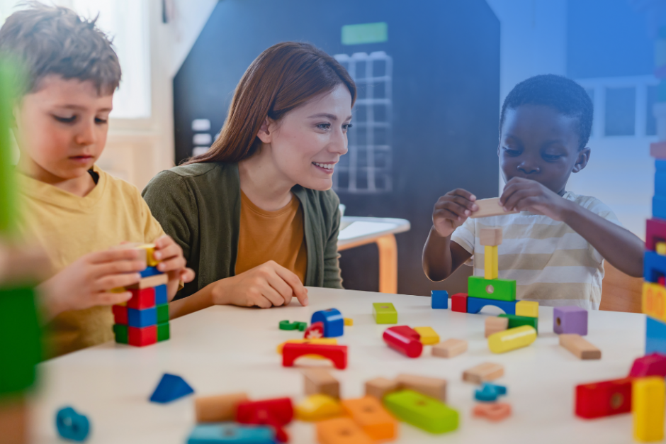 A teacher smiles while two children build colorful block towers at a table in a preschool classroom.