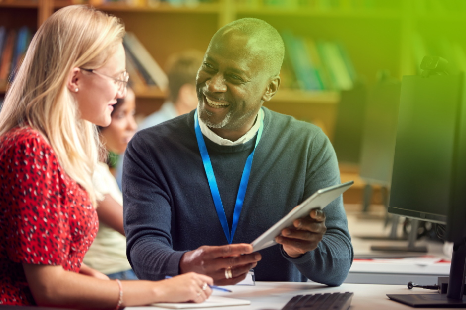 A principal in a navy blue sweater and lanyard smiles while discussing something on a tablet with a teacher in a red blouse. They are in a library setting.