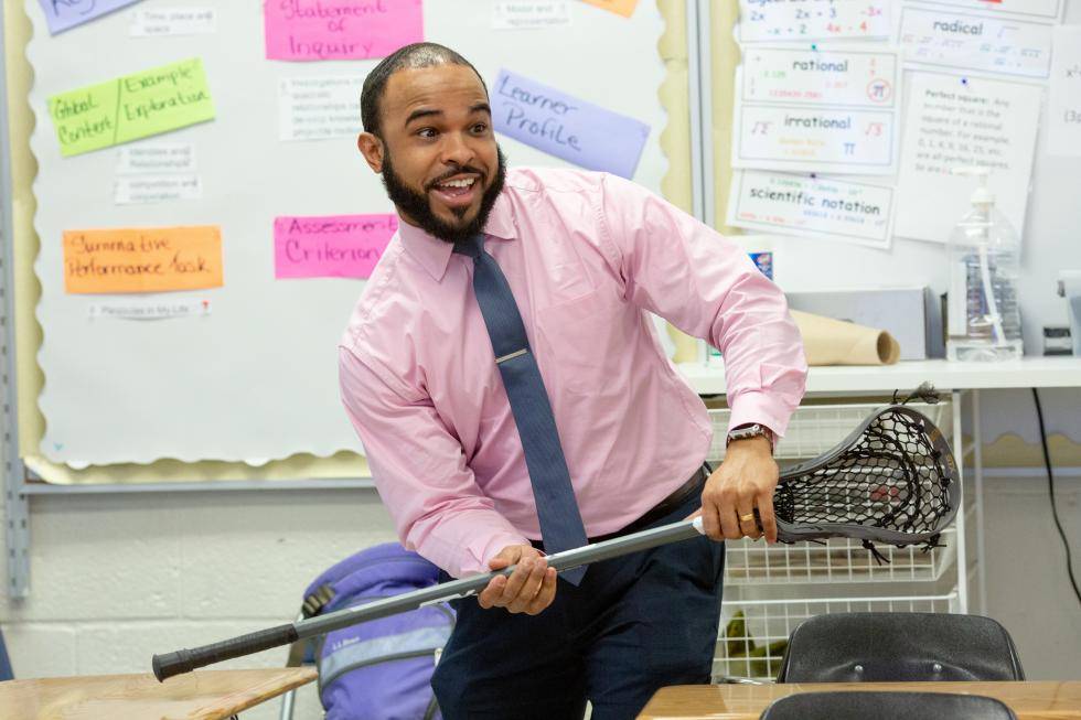 male teacher smiling, holding a lacrosse stick at the front of the class