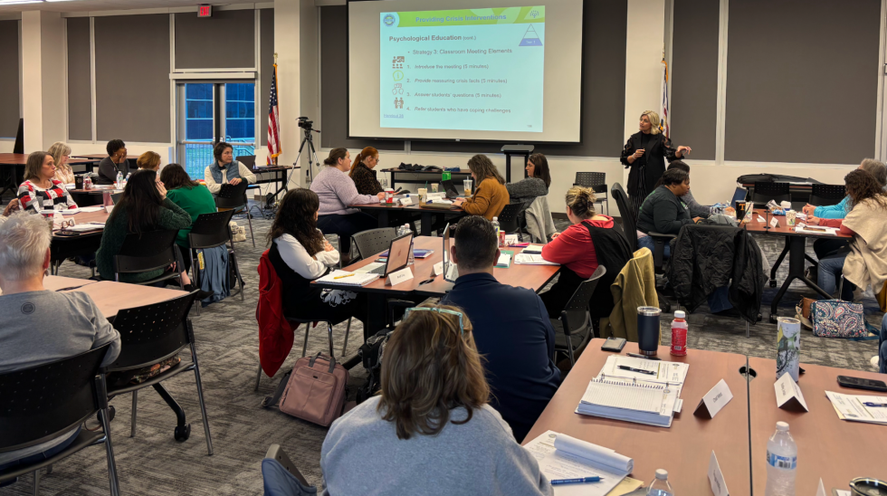 people sitting at desks looking at a screen with a person standing in the front of the room