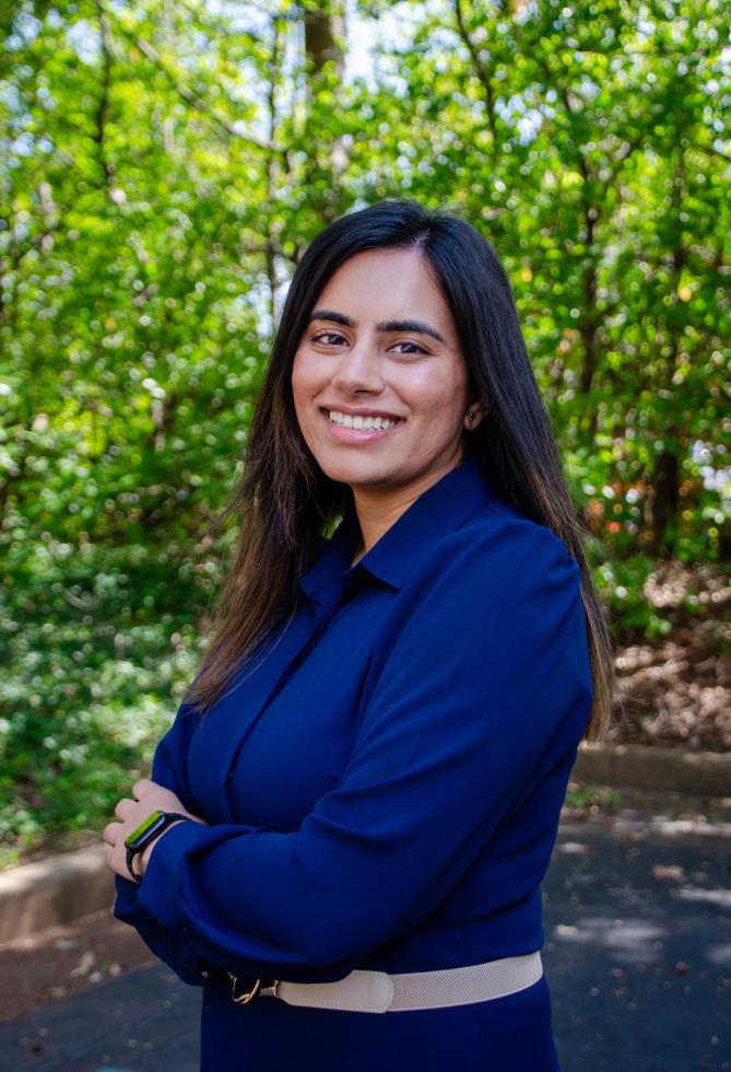 Raheela Charania stands smiling with her arms crosses in front of some green trees. 