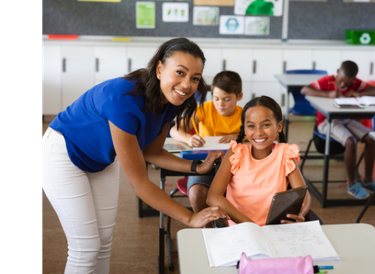 A teacher in a blue shirt supports a smiling student who is using a tablet and has a workbook open on their desk.  Two more students are in the background, focused on their papers in a bright, colorful classroom.