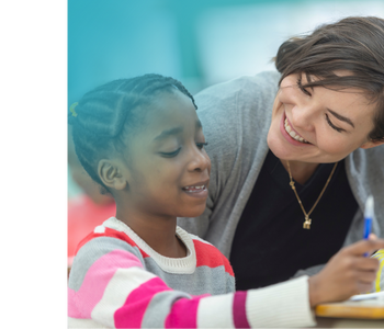 A teacher smiling at an elementary school student who is sitting at her desk.
