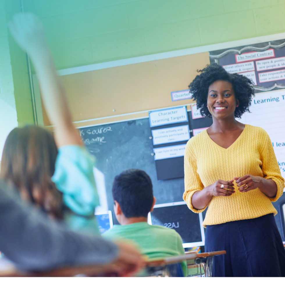 A teacher standing at the front of the classroom smiling while a student sitting at a desk raises their hand.
