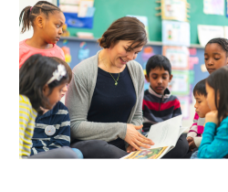A teacher sitting on the floor reading a book surrounded by young students